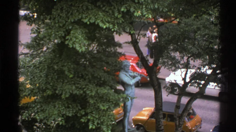 End frame from a 1976 Norway 8mm clip showing a bronze street statue among leafy trees with cars and pedestrians nearby.