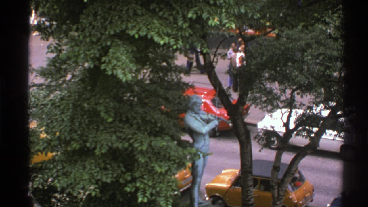 1976 Norway 8mm frame showing a leafy street, blue-green violinist statue, parked red and yellow cars, and pedestrians in the background.