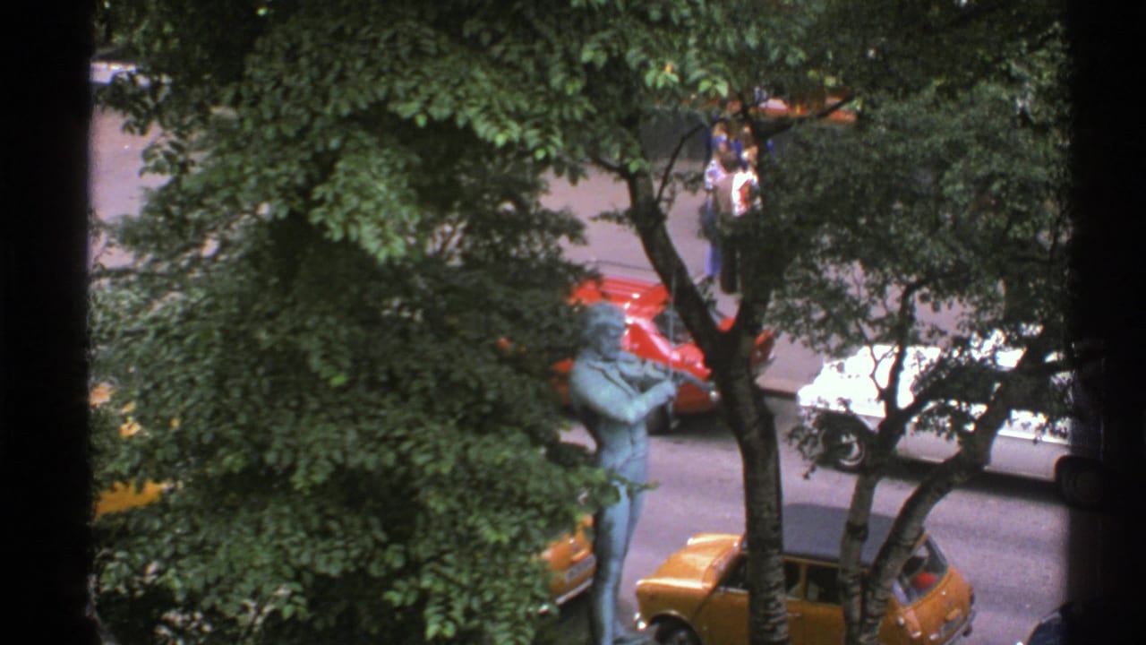 1976 Norway street view from a window, with dense trees, a blue-green statue, parked cars, and distant pedestrians near the sidewalk.