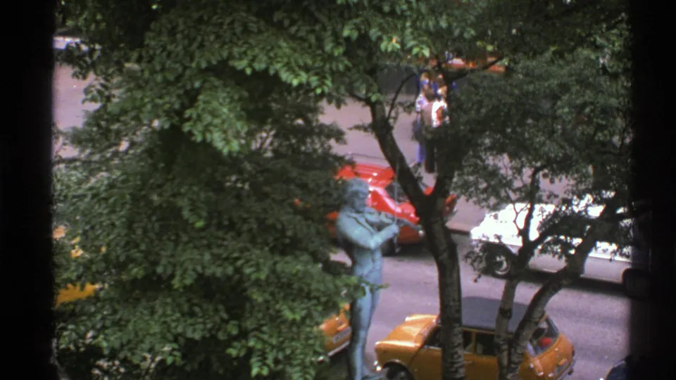 1976 Norway street view from a window, with dense trees, a blue-green statue, parked cars, and distant pedestrians near the sidewalk.