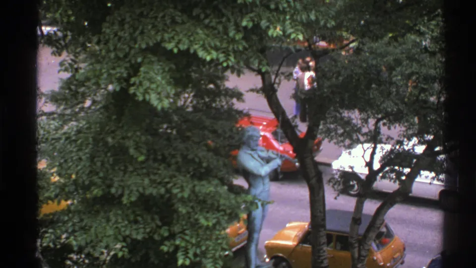 1976 Norway 8mm frame of a blue-green statue near leafy trees, with parked yellow, red, and white cars and pedestrians beyond.