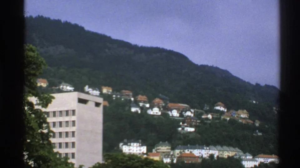 1976 Norway view of hillside housing beneath a wooded mountain, with a pale rectangular building in front.
