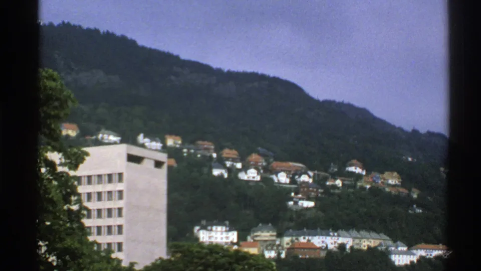 1976 Norway frame with the camera shifted lower, showing hillside houses and the edge of a pale block building.