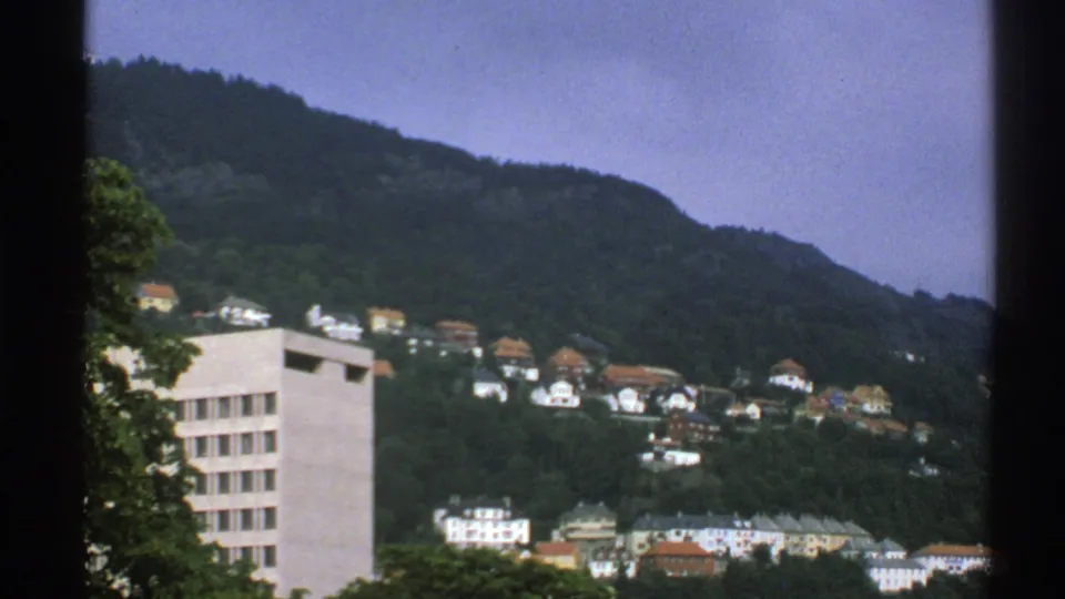 1976 Norway home-movie frame of hillside residences beneath a wooded ridge, with dark frame edges visible.