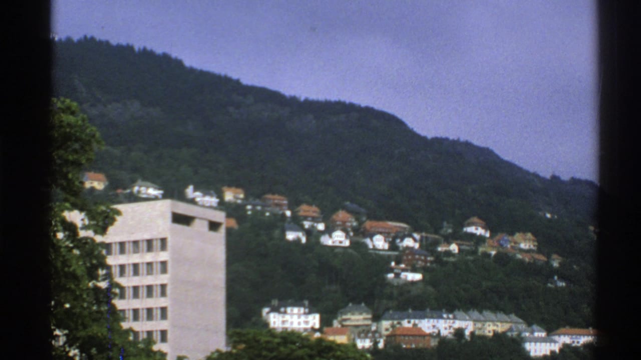 1976 Norway 8mm view of hillside homes among trees, with a pale modern building in the foreground and dark frame edges.