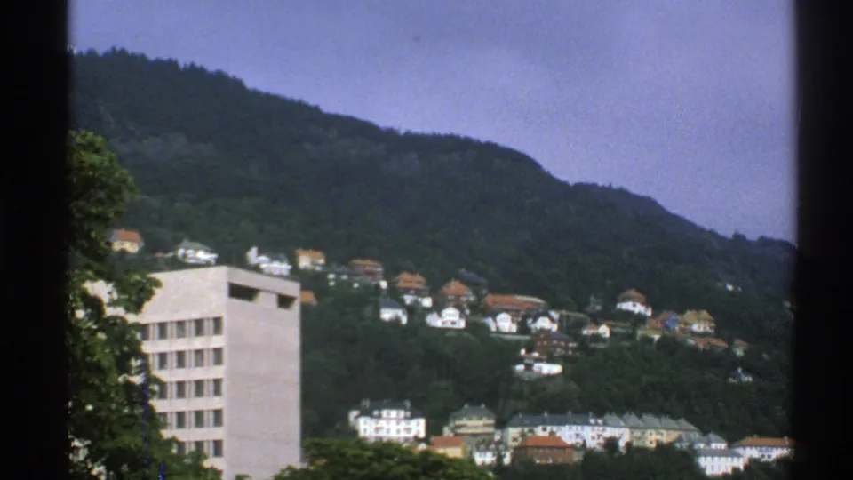 1976 Norway 8mm view of hillside homes among trees, with a pale modern building in the foreground and dark frame edges.