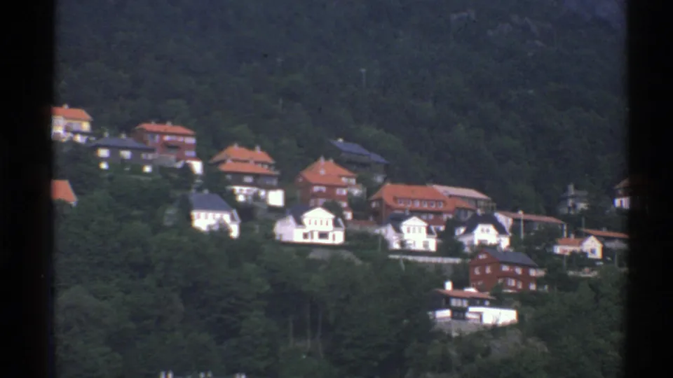 1976 Norway home-movie frame of hillside residences and forested slopes, divided by a dark window or film-frame edge.