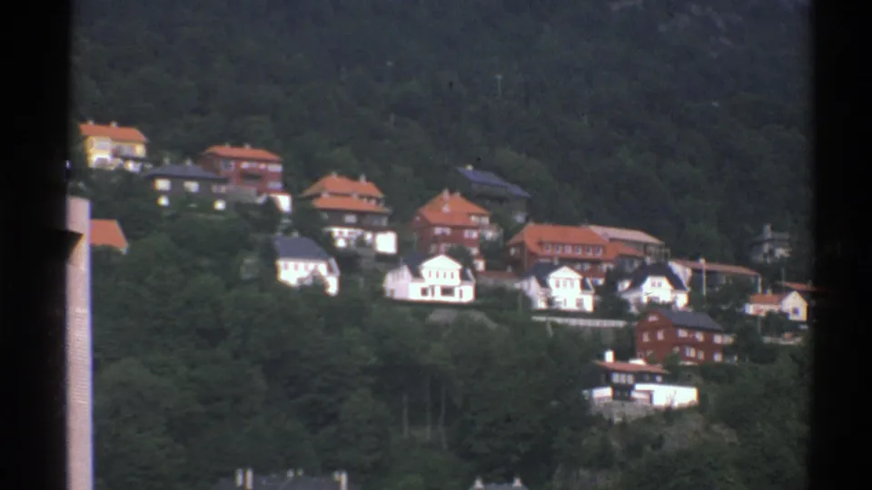 1976 Norway home-movie frame of a wooded hillside village with white homes, red roofs, and a few darker structures.
