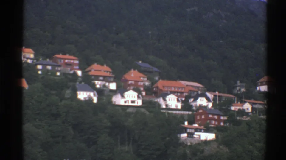 1976 Norway home-movie frame of hillside houses, red roofs, and white walls partly screened by trees.