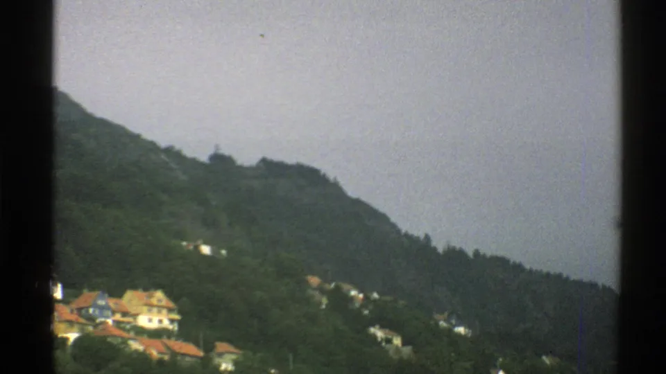 Norway, 1976: distant red-roofed homes sit near the lower edge of a forested hillside under grey cloud cover.