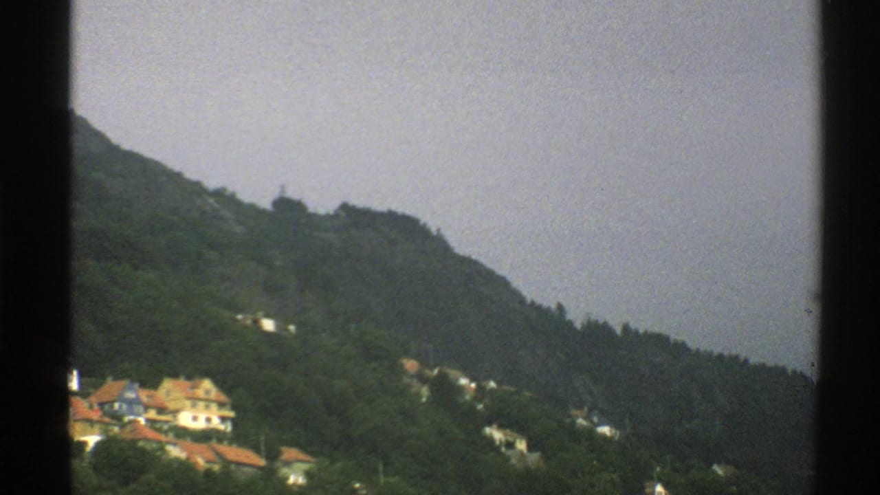 Norway 1976 home-movie frame showing hillside houses and a green mountain, with airport activity partly out of view.