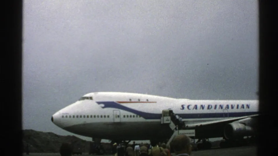 Norway 1976 frame of a Scandinavian Airlines Boeing 747 on the tarmac, with passengers gathered around mobile stairs near the fuselage.