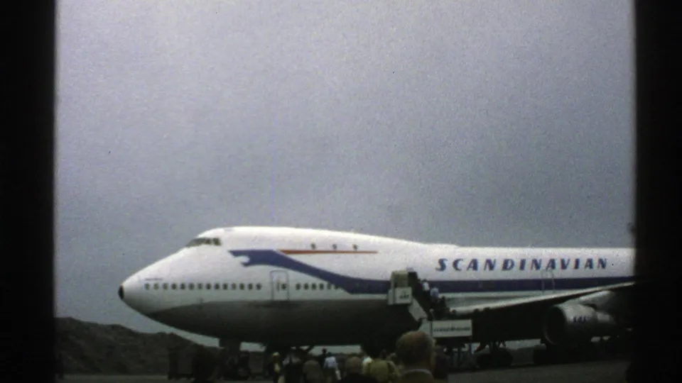 1976 Norway 8mm frame showing a Scandinavian Airlines 747 at a small airport with boarding stairs and passengers near the fuselage.