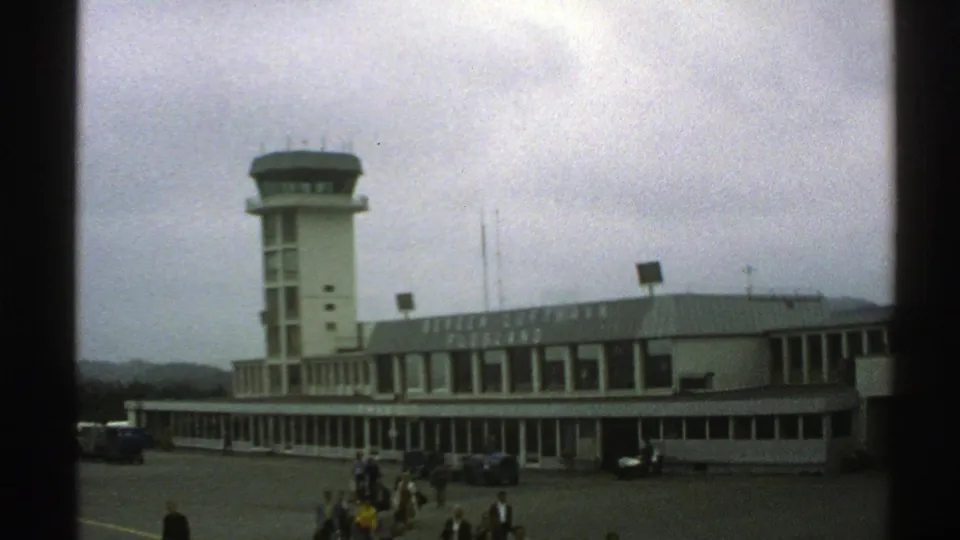 Norway 1976 airport terminal view with a modest control tower, low building, parked cars, and travelers outside.