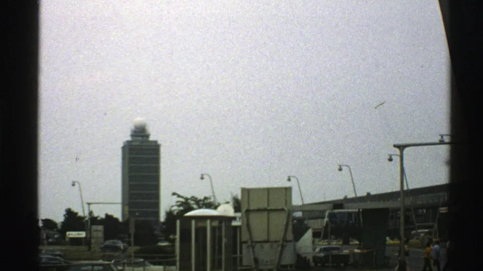 1976 8mm frame showing a distant tower beyond cars, streetlights, signage, and small figures near a roadway.