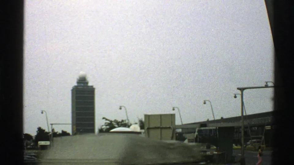 1976 8mm frame with motion blur from a passing vehicle crossing the view of a distant tall building.