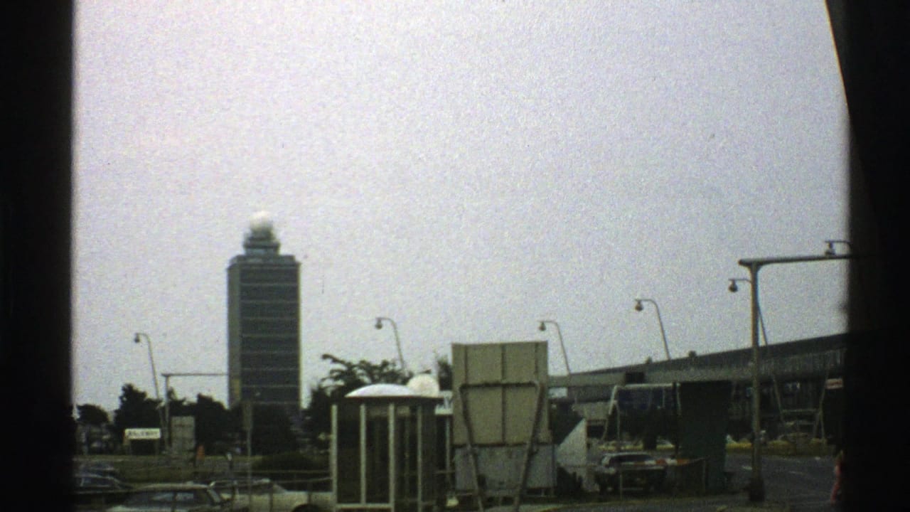 1976 Norway 8mm frame showing a tall control-tower-like building beyond streetlights, cars, and roadside structures.