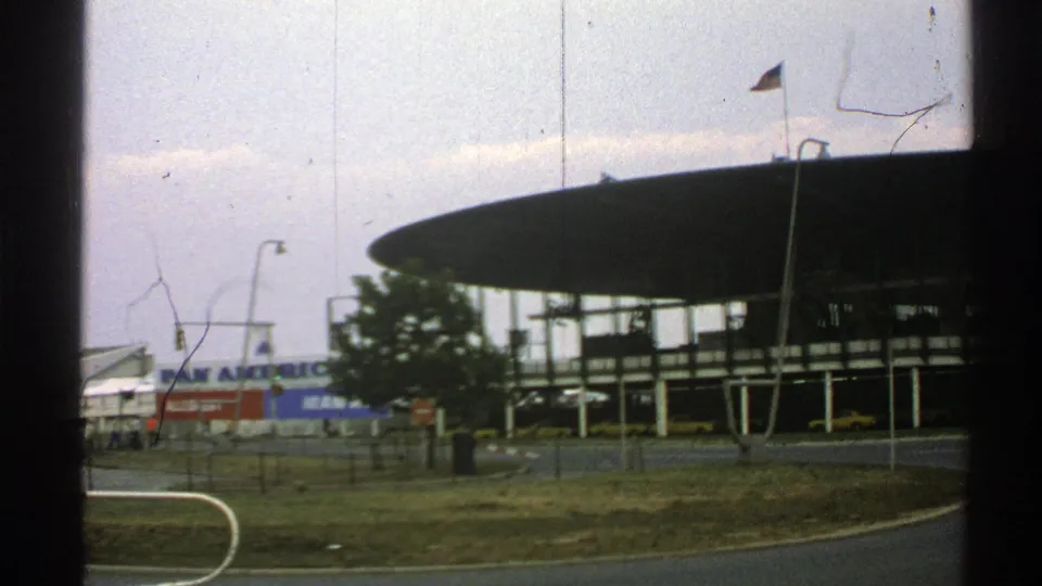 1976 Norway 8mm frame of the airport terminal through a vehicle window, with Pan Am signage partly visible and film scratches across the sky.