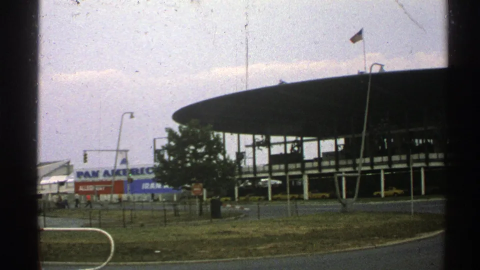 1976 Norway home-movie frame showing a modernist airport terminal, Pan Am sign, flagpole, road edge, grass verge, and dark window border.