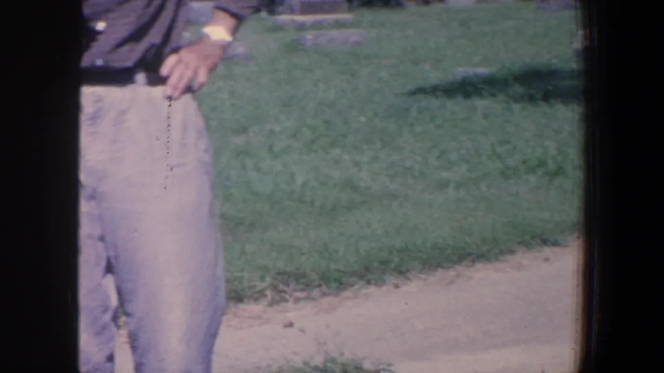 Cropped 1959 Iowa cemetery frame showing a man in light trousers with grass and walkway behind him.