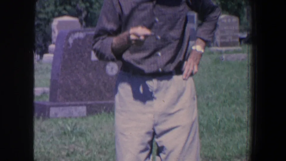 1959 Iowa 8mm frame of a man standing in a grassy cemetery with gravestones visible behind him.