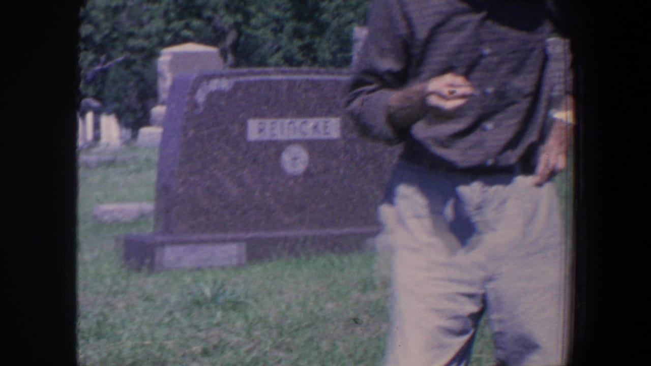 In 1959 Iowa home-movie footage, a man in light trousers passes a REINCKE gravestone in a sunlit grassy cemetery.