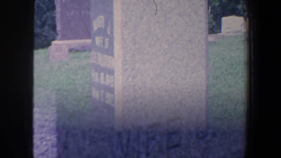 The 8mm camera moves close to a weathered cemetery marker, with partial lettering visible and grass behind it.