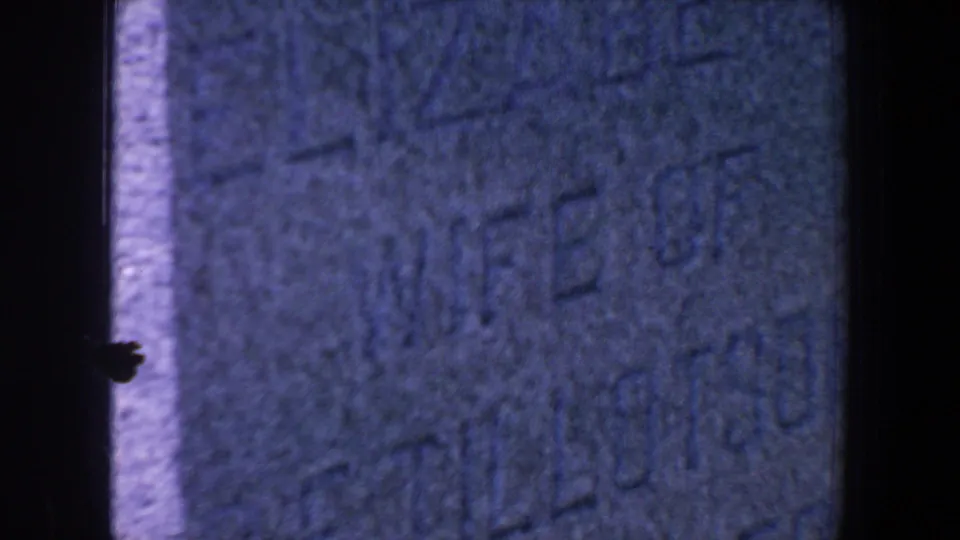 Close 1959 Iowa 8mm view of a worn gravestone face with partial carved text and dark film edges.