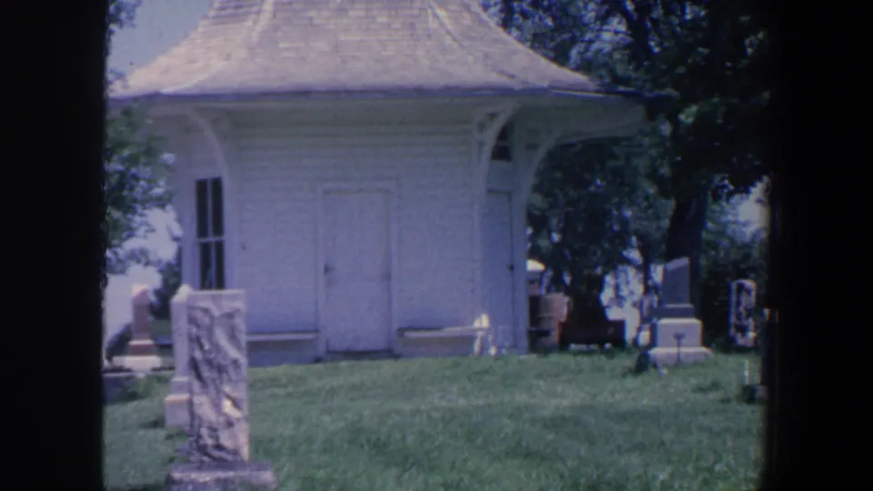1959 Iowa cemetery view showing a small white building, shingled roof, lawn, trees, and upright stones.