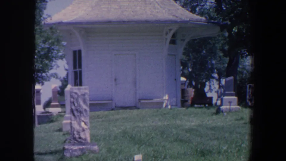 1959 Iowa 8mm frame of a white cemetery structure, foreground grave marker, grass, trees, and surrounding stones.