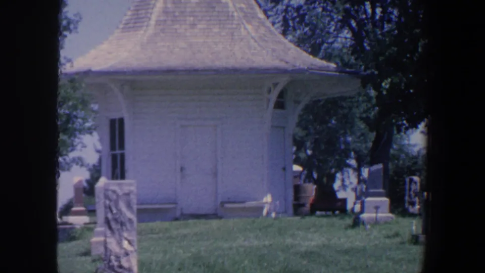 1959 Iowa cemetery frame with the white building, roofline, grave markers, grass, trees, and dark 8mm gate borders.