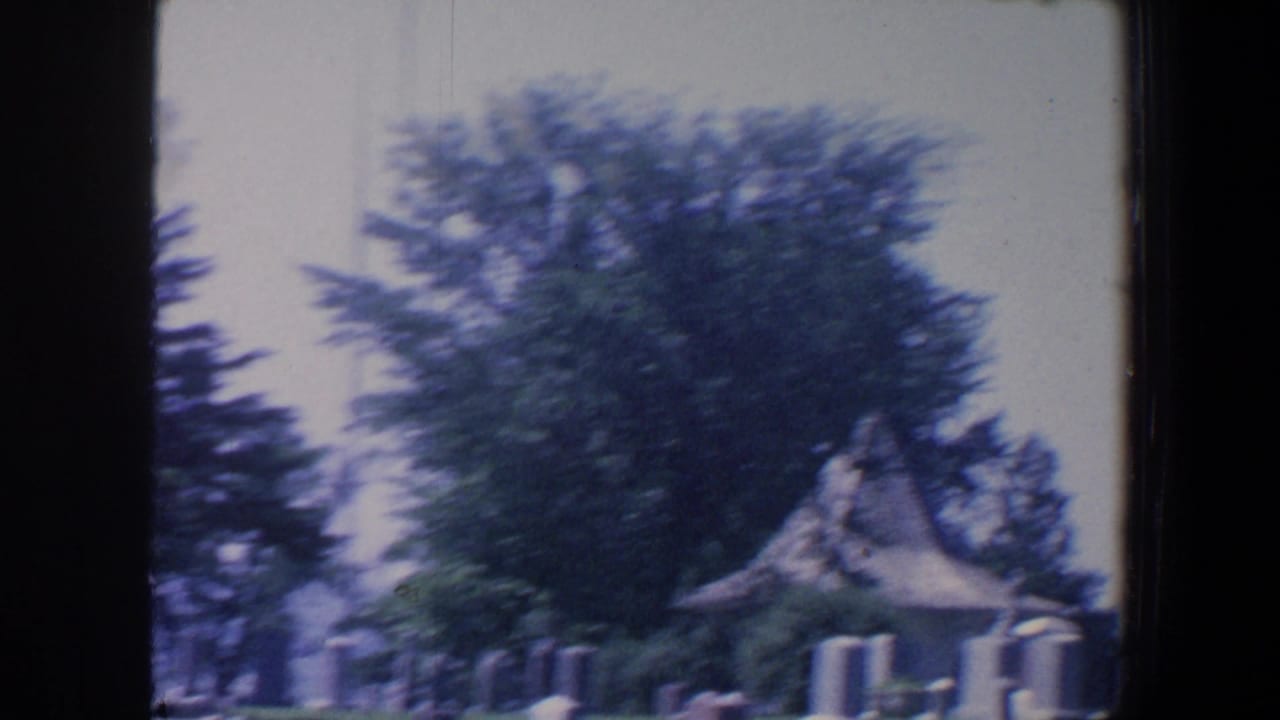 1959 Iowa 8mm frame showing a cemetery lawn with grave markers, trees, and a small chapel-like shelter in soft focus.
