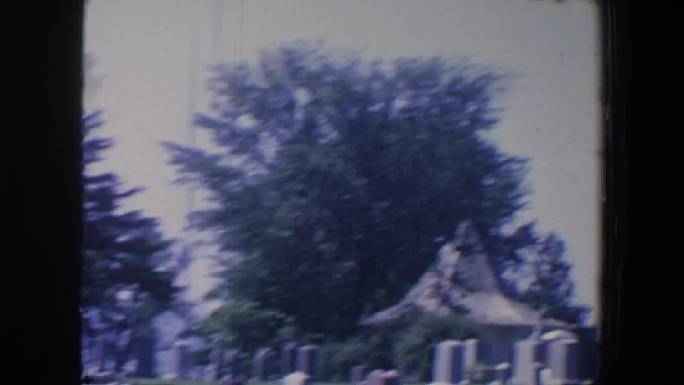 1959 Iowa 8mm frame showing a cemetery lawn with grave markers, trees, and a small chapel-like shelter in soft focus.