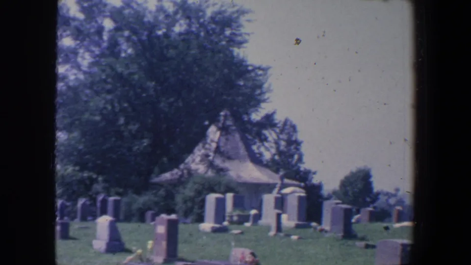 1959 Iowa cemetery grounds with upright grave markers, floral tributes, mature trees, and a small chapel-like structure.