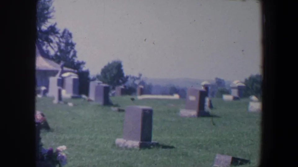 1959 Iowa 8mm frame looking across a cemetery lawn toward scattered tombstones, flowers, trees, and a distant horizon.