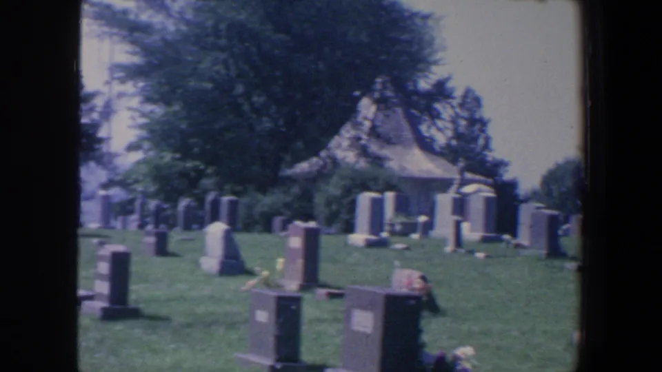 1959 Iowa cemetery scene with foreground grave markers, floral decorations, trees, and a small peaked-roof shelter behind them.