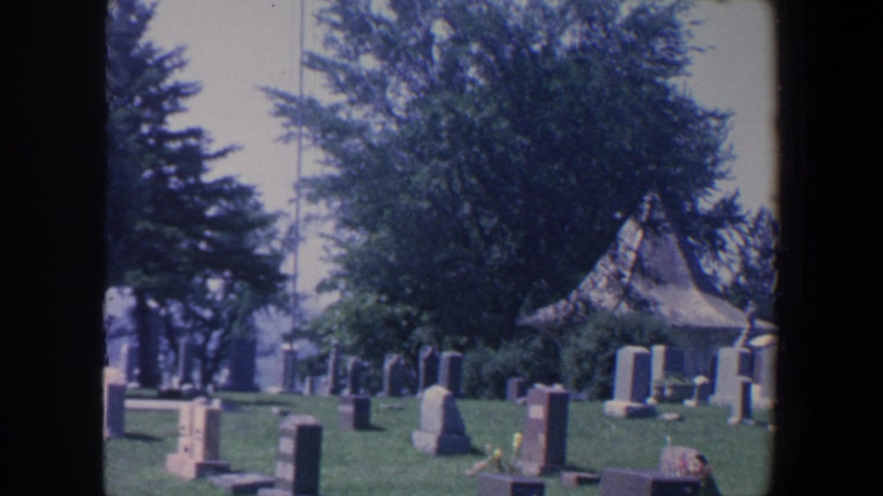 1959 Iowa 8mm frame showing a quiet cemetery with green grass, upright headstones, mature trees, and a small chapel-like structure.