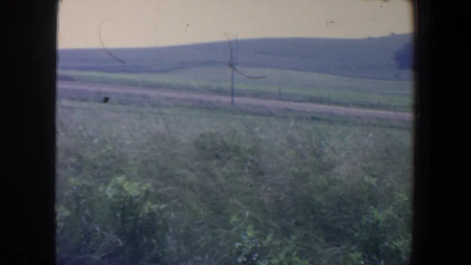 1959 Iowa 8mm frame looking over green rural fields, a dirt road, fence line, and low rolling land under a pale sky.