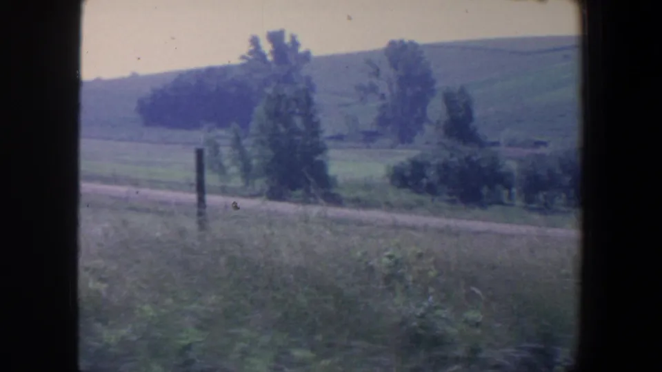 1959 Iowa rural landscape with green hills, scattered trees, roadside grass, and a dirt or gravel road in soft 8mm color.