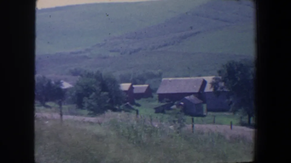1959 Iowa 8mm frame showing farm buildings, trees, grass, and rolling hills from a distant roadside viewpoint.