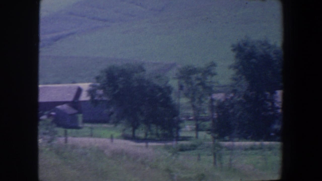 Farmstead with low buildings, trees, and grassy fields beneath distant hills.