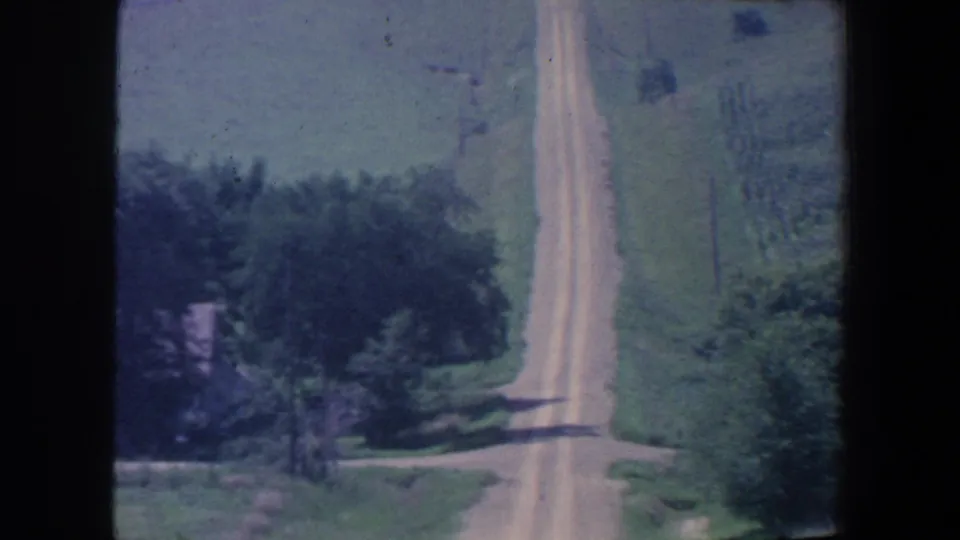 1959 Iowa 8mm frame showing a narrow rural road bordered by trees, grass, and open farmland.