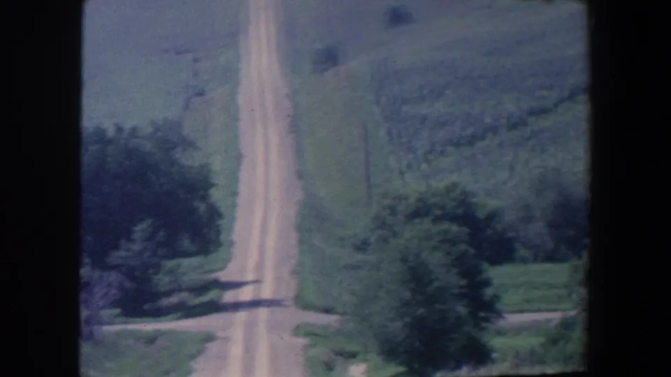 1959 Iowa 8mm frame showing a pale dirt road through rolling green fields, trees, and utility poles.