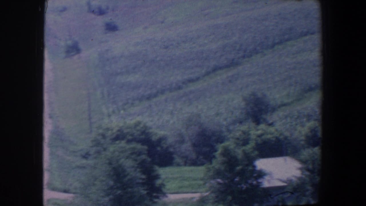 Rolling fields and trees with a small light-roofed farm structure at lower right.
