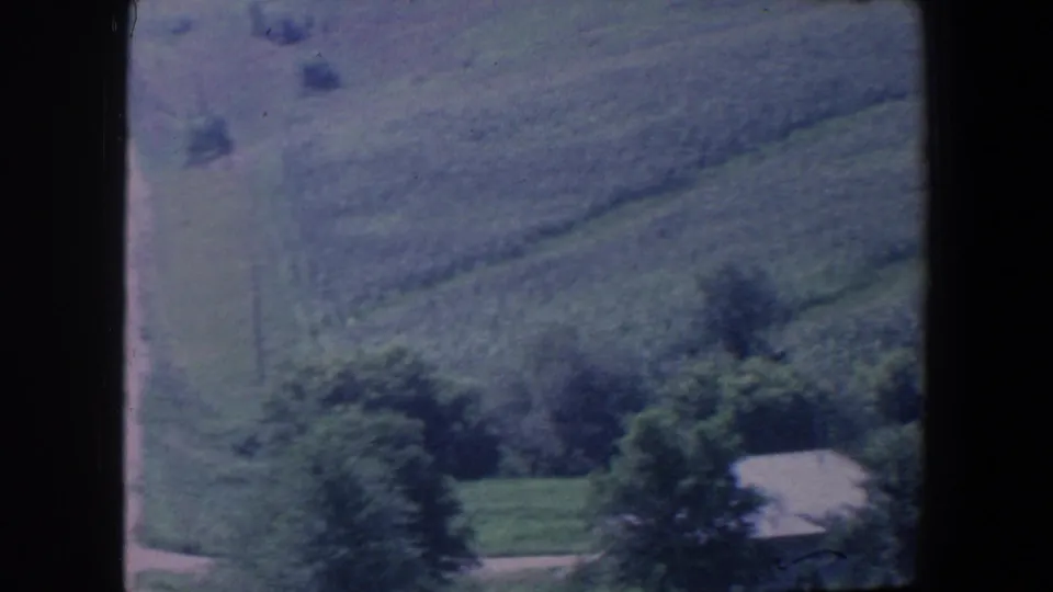1959 Iowa countryside frame with rolling green fields, dense trees, and a small light-roofed farm structure.