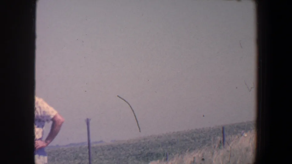 1959 Iowa home-movie frame with a partially visible adult man in a patterned shirt near a field and fence posts under a pale sky.