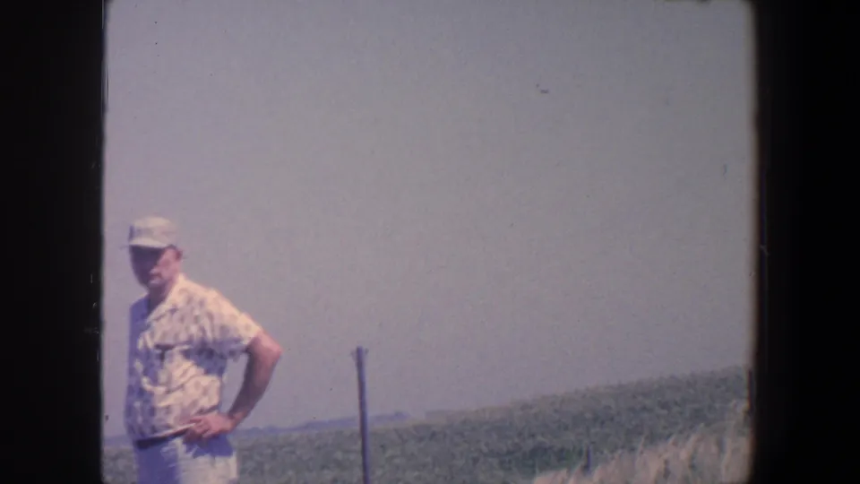An adult man in a cap and patterned short-sleeve shirt stands hands on hips in a 1959 Iowa field landscape.