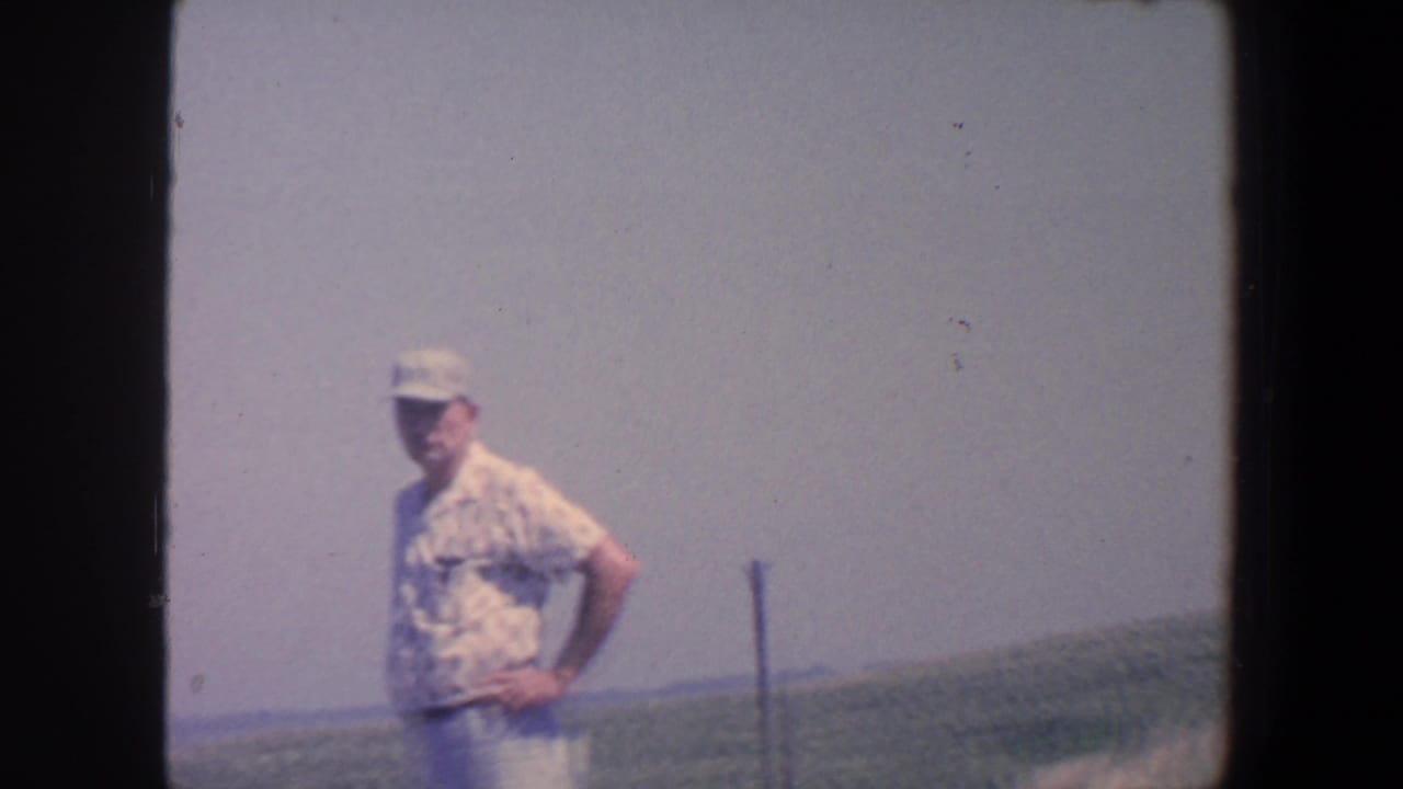 Adult man in 1959 Iowa stands with hands on hips beside open farmland and a distant horizon.