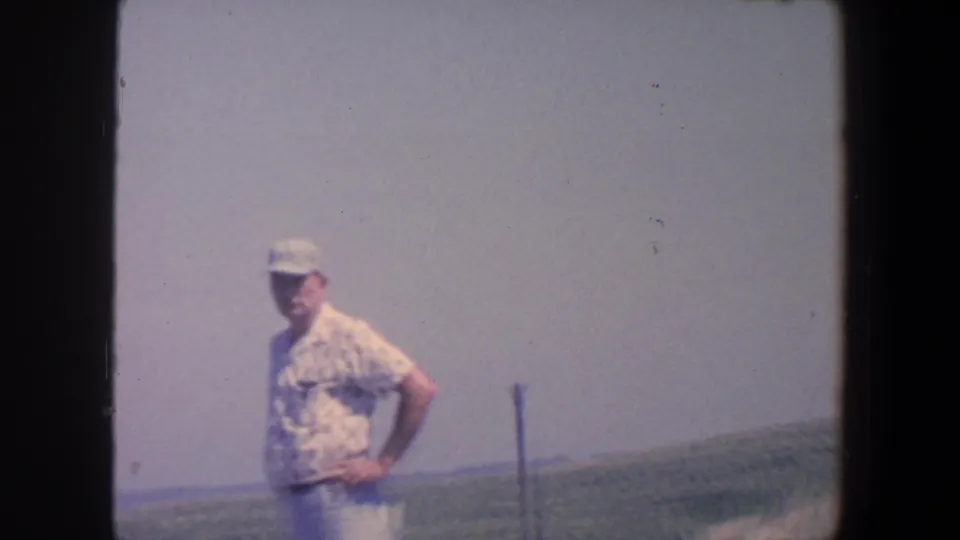 Adult man in 1959 Iowa stands with hands on hips beside open farmland and a distant horizon.
