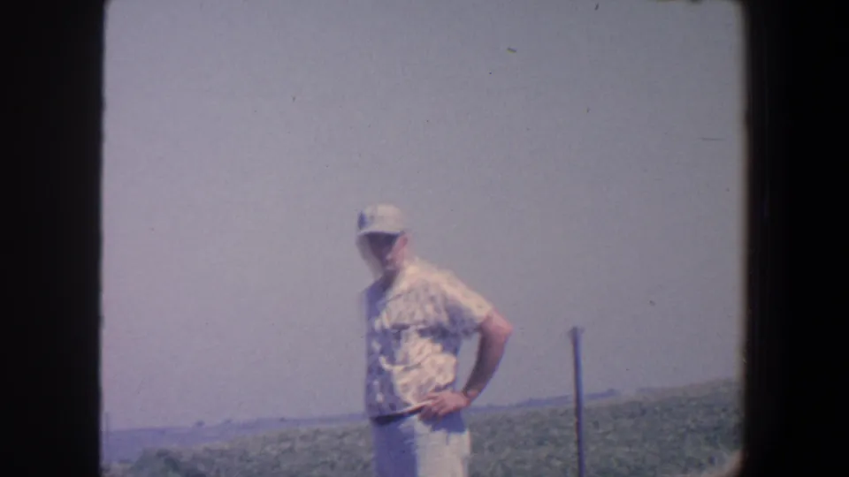 Adult man in 1959 Iowa turns slightly while standing near rolling farmland beneath a pale sky.